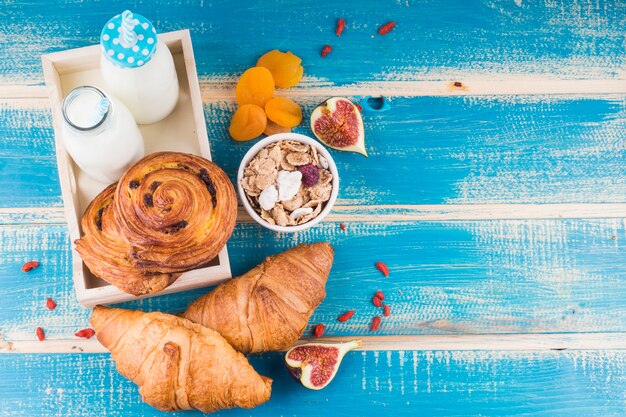 Baked breads with milk bottles in tray near dry apricots; fig fruit; and corn flakes over blue wooden background