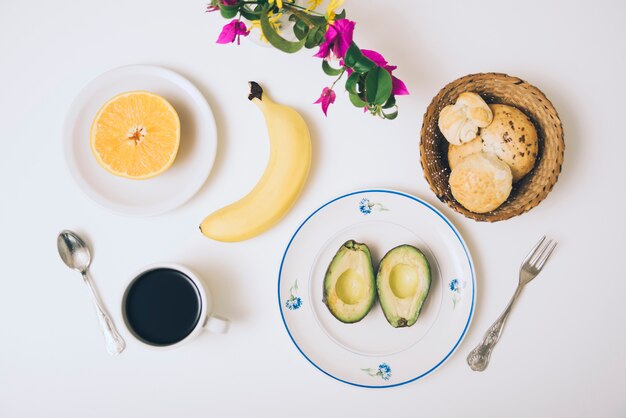 Baked buns; avocado; banana; halved orange; coffee and flowers on white backdrop