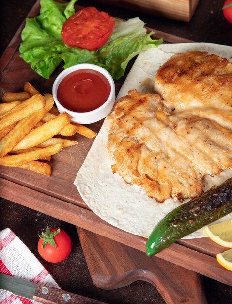 Baked chicken breast with French fries in lavash with vegetables and ketchup on wooden board 