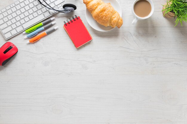Baked croissant and tea cup with keyboard and office supplies on wooden table with space for writing text