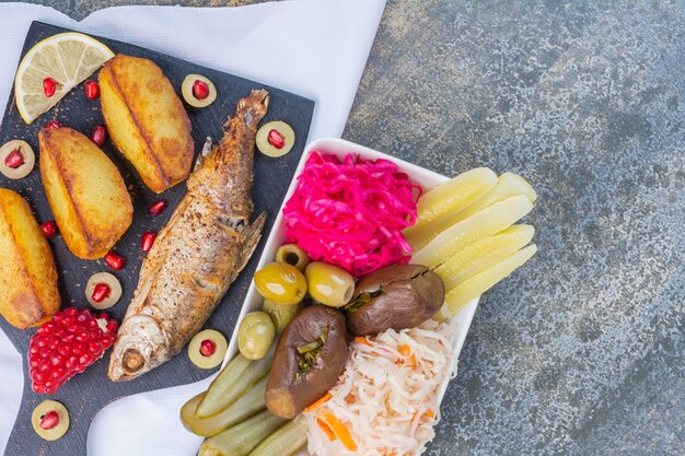 Baked fish and fried potato on a cutting board next to a bowl of preserved vegetables.