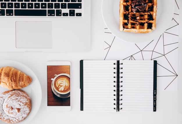 Запеченная еда; шоколадные вафли; мобильный телефон с кофейным экраном; ноутбук и спиральный блокнот на белом столе