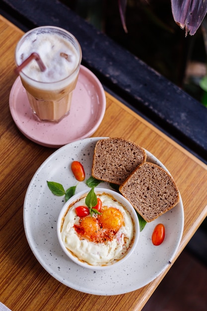 Запеченные два яйца с ветчиной и сыром в круглой миске два кусочка черного хлеба сбоку