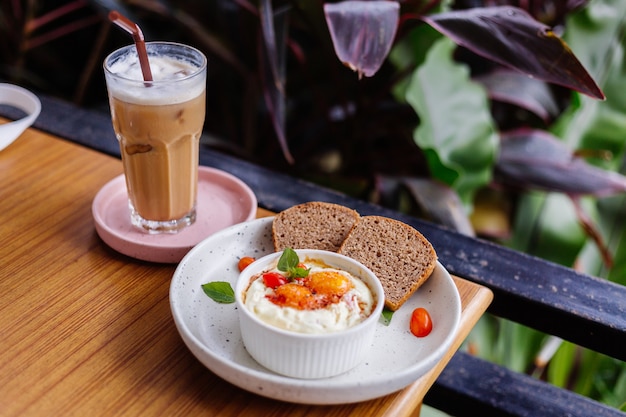 Запеченные два яйца с ветчиной и сыром в круглой миске два кусочка черного хлеба сбоку