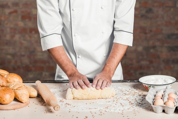 Baker kneading dough on kitchen worktop with baked breads and ingredients