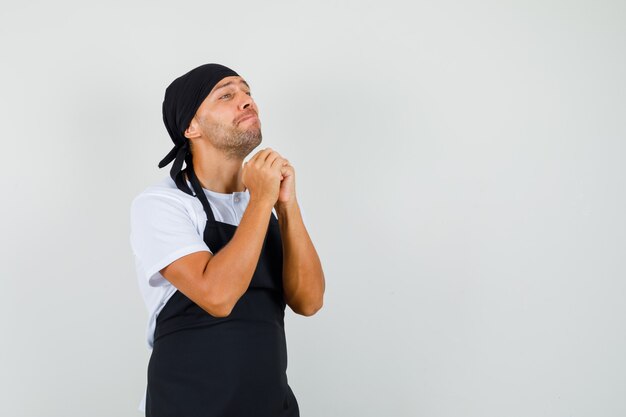 Baker man in t-shirt, apron clasping hands in praying gesture and looking helpless