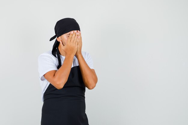 Baker man in t-shirt, apron covering face with hands and looking depressed