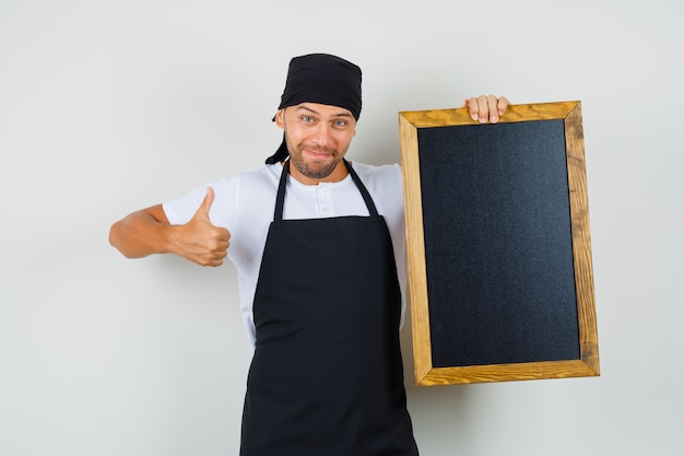 Baker man in t-shirt, apron holding blackboard, showing thumb up and looking happy