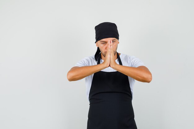 Baker man in t-shirt, apron holding hands in praying gesture and looking calm