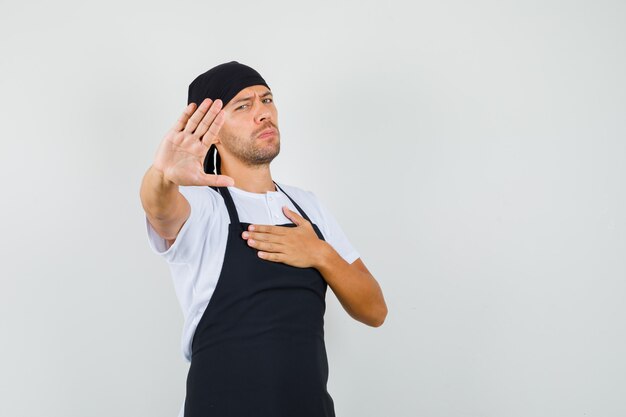 Baker man in t-shirt, apron showing stop gesture with hand on chest and looking bored