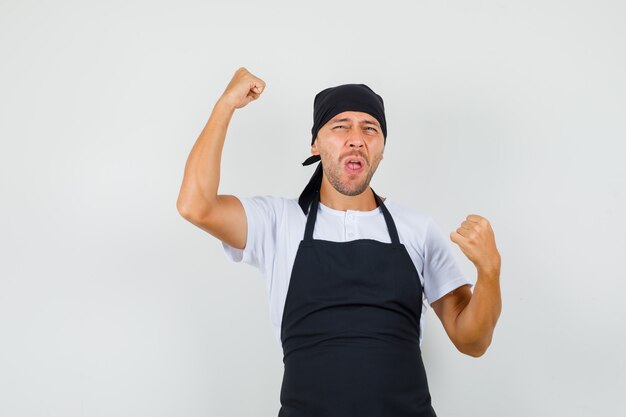 Мужчина-пекарь в футболке, фартуке показывает жест победителя и выглядит счастливым
