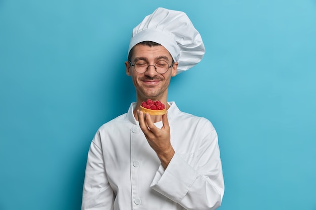 Bakery chef smells handmade freshy baked cakes decorated with raspberry, works at pastry shop, closes eyes in pleasure