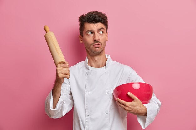 Bakery and cooking concept. Pensive surprised professional cook holds wooden rolling pin and bowl