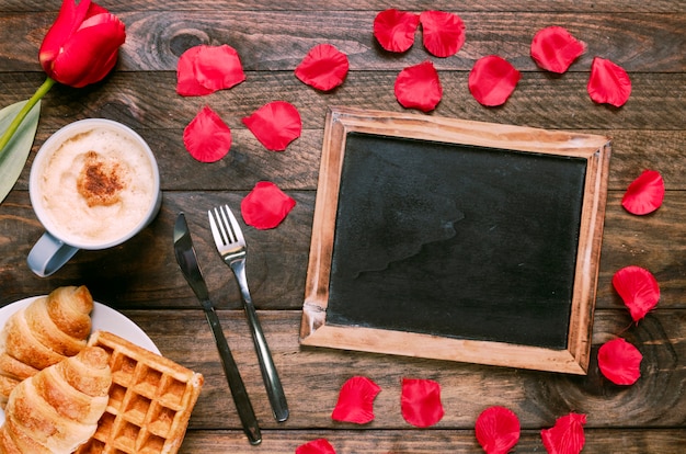 Bakery on plate near cup of drink, flower, cutlery, petals and photo frame