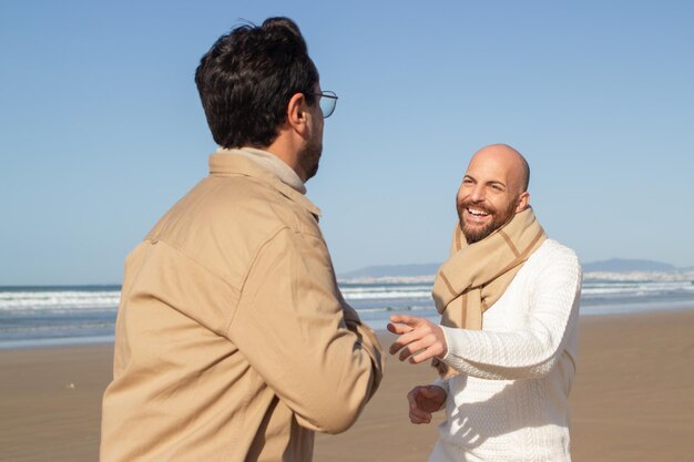 Bald gay man laughing while walking with boyfriend. Positive middle-aged homosexual couple walking together on beach. Dating concept