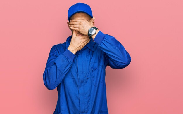 Bald man with beard wearing builder jumpsuit uniform covering eyes and mouth with hands, surprised and shocked. hiding emotion