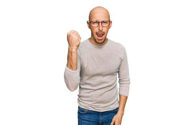 Bald man with beard wearing casual clothes and glasses angry and mad raising fist frustrated and furious while shouting with anger rage and aggressive concept