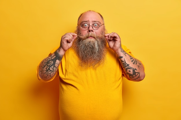 Free photo bald surprised man curls mustache, has thick beard, stares with disbelief, wears transparent glasses, dressed in casual clothes, has fat belly poses indoor. fatso male poses with astonished expression