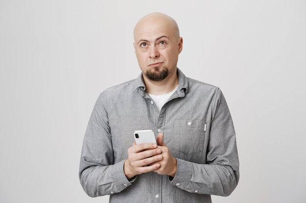 Бесплатное фото Лысый думающий парень смотрит вверх, держа мобильный телефон