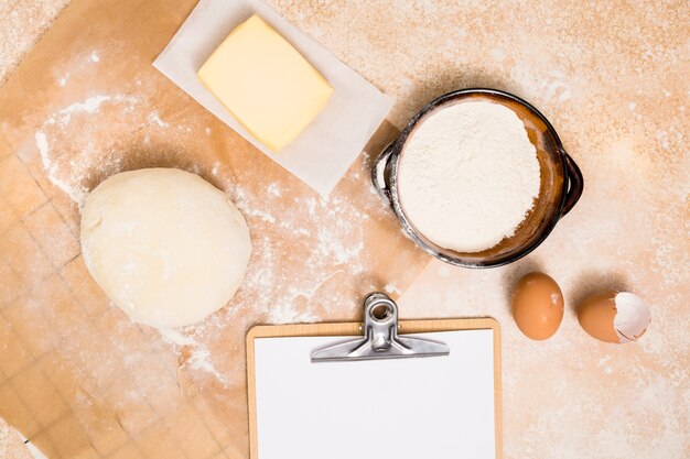 Ball of dough; flour; block of butter; eggs and clipboard over kitchen backdrop