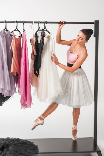 Ballerina standing on tiptoe choosing tutu from hangers against white backdrop