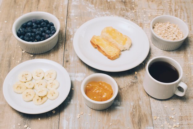 Banana slices; jam; blueberries; oats and coffee cup on wooden table