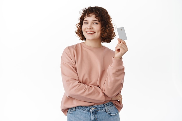 Free photo bank and finance concept. confident and satisfied young woman shows credit card, smiles happy and pleased, paying contactless, going shopping, standing over white background