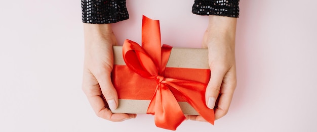 Banner made of woman hands holding present box with red bow on pink background flat lay top view