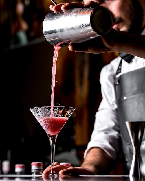Бар тендер наливает коктейль из коктейльного шейкера в бокал для мартини