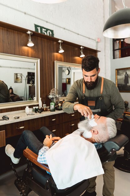 Парикмахер наносит крем для бритья пожилому мужчине в парикмахерской