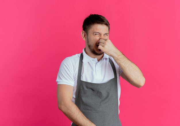 barber man in apron closing nose with fingers suffering from stench standing over pink wall