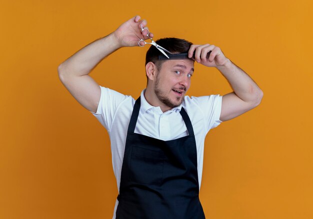 Barber man in apron combing and cutting his hair standing over orange background