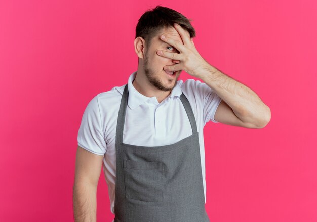Barber man in apron covering eyes with fingers looking through fingers smiling standing over pink background