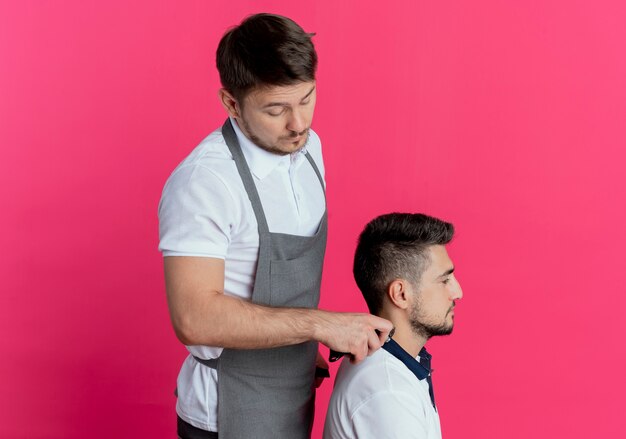 barber man in apron cutting hair with hair cutting machine of satisfied client over pink wall