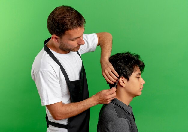 緑の壁の上に立っている満足しているクライアントのはさみで髪を切るエプロンの床屋の男