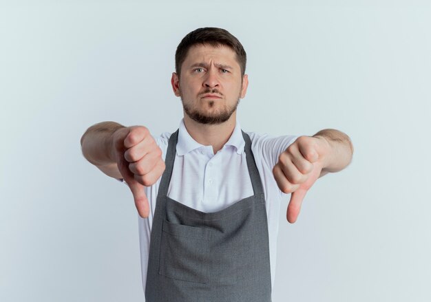 barber man in apron  displeased showing thumbs down standing over white wall