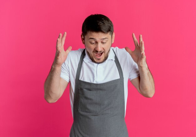 barber man in apron going wild shouting with arms raised standing over pink wall