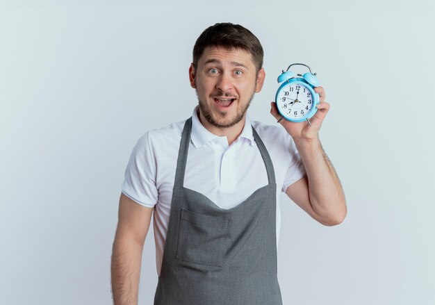 Barber man in apron holding alarm clock looking at camera happy and surprised standing over white background