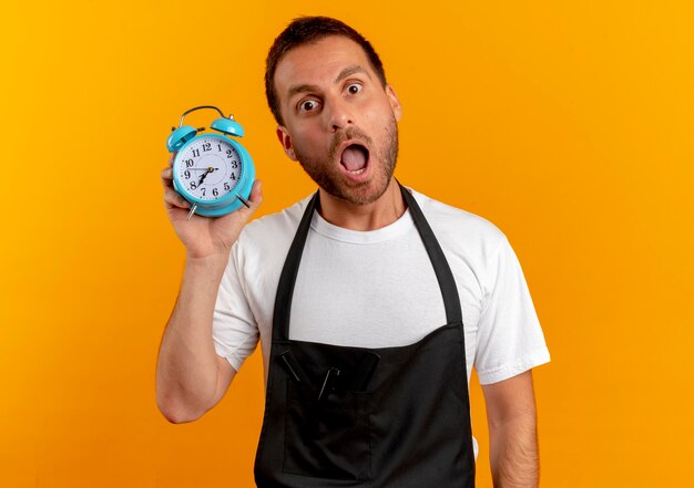 Free photo barber man in apron holding alarm clock looking to the front worried with wide open mouth standing over orange wall