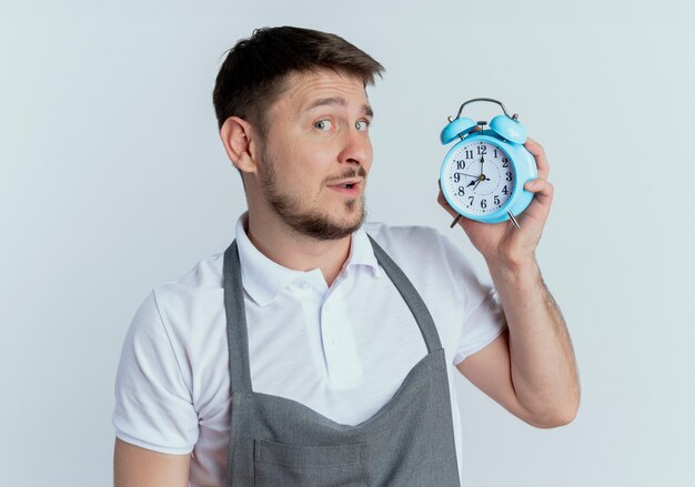 Barber man in apron holding alarm clocklooking at camera with skeptic smile on face standing over white background