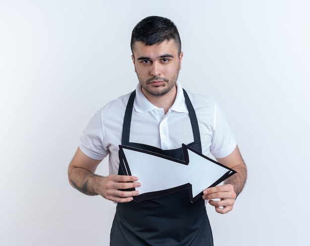 Barber man in apron holding arrow looking at camera with serious confident expression standing over white background