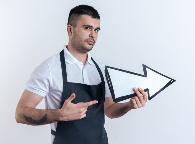 Barber man in apron holding arrow pointing with index finger at it looking confident standing over white