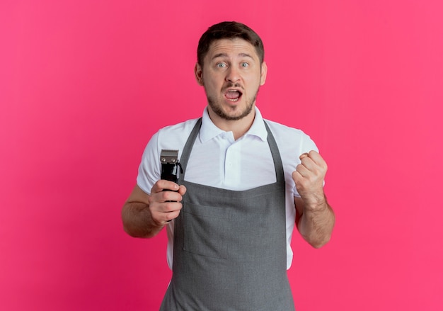 Barber man in apron holding beard trimmer clenching fist happy and excited standing over pink background