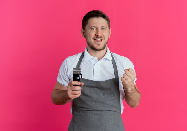 Barber man in apron holding beard trimmer clenching fist happy and excited standing over pink background