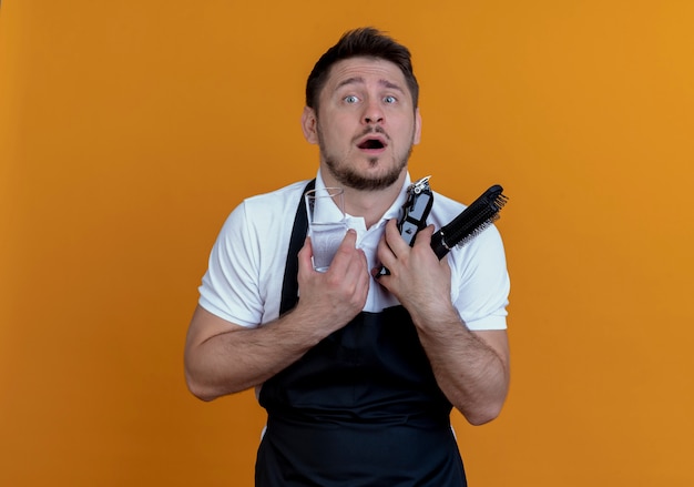 Barber man in apron holding beard trimmer, hair brush and glass of water looking at camera confused standing over orange background