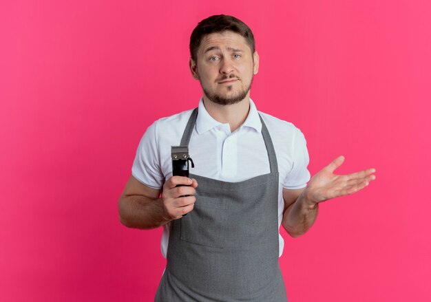 Barber man in apron holding beard trimmer looking at camera confused and displeased standing over pink background
