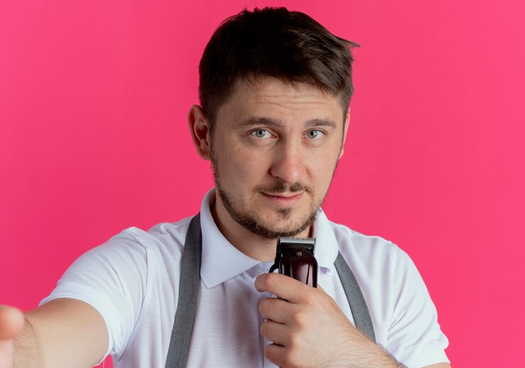 मुफ़्त फोटो barber man in apron holding beard trimmer looking at camera with smile on face standing over pink background