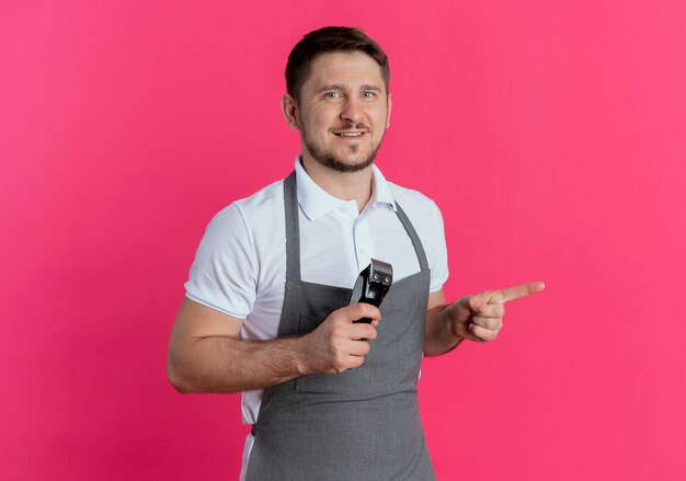 парикмахер в фартуке держит триммер для бороды, указывая пальцем в сторону, улыбаясь, стоя над розовой стеной