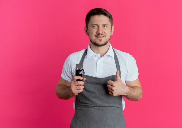 ピンクの背景の上に立っているカメラを見て笑顔で親指を示すひげトリマーを保持しているエプロンの理髪店の男