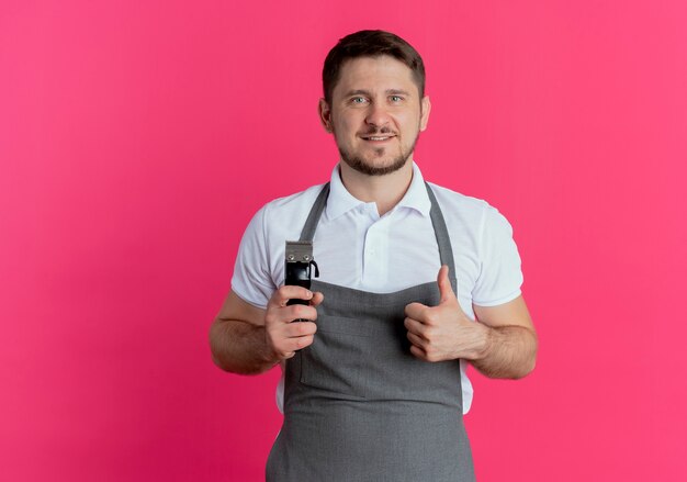 Barber man in apron holding beard trimmer showing thumbs up smiling looking at camera standing over pink background